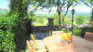 Une table en bois placée à l’extérieur, entourée de chaises, avec un saladier rempli de citrons et une bouteille de jus d’orange, le tout mis en valeur par un décor de verdure et un mur de pierres.