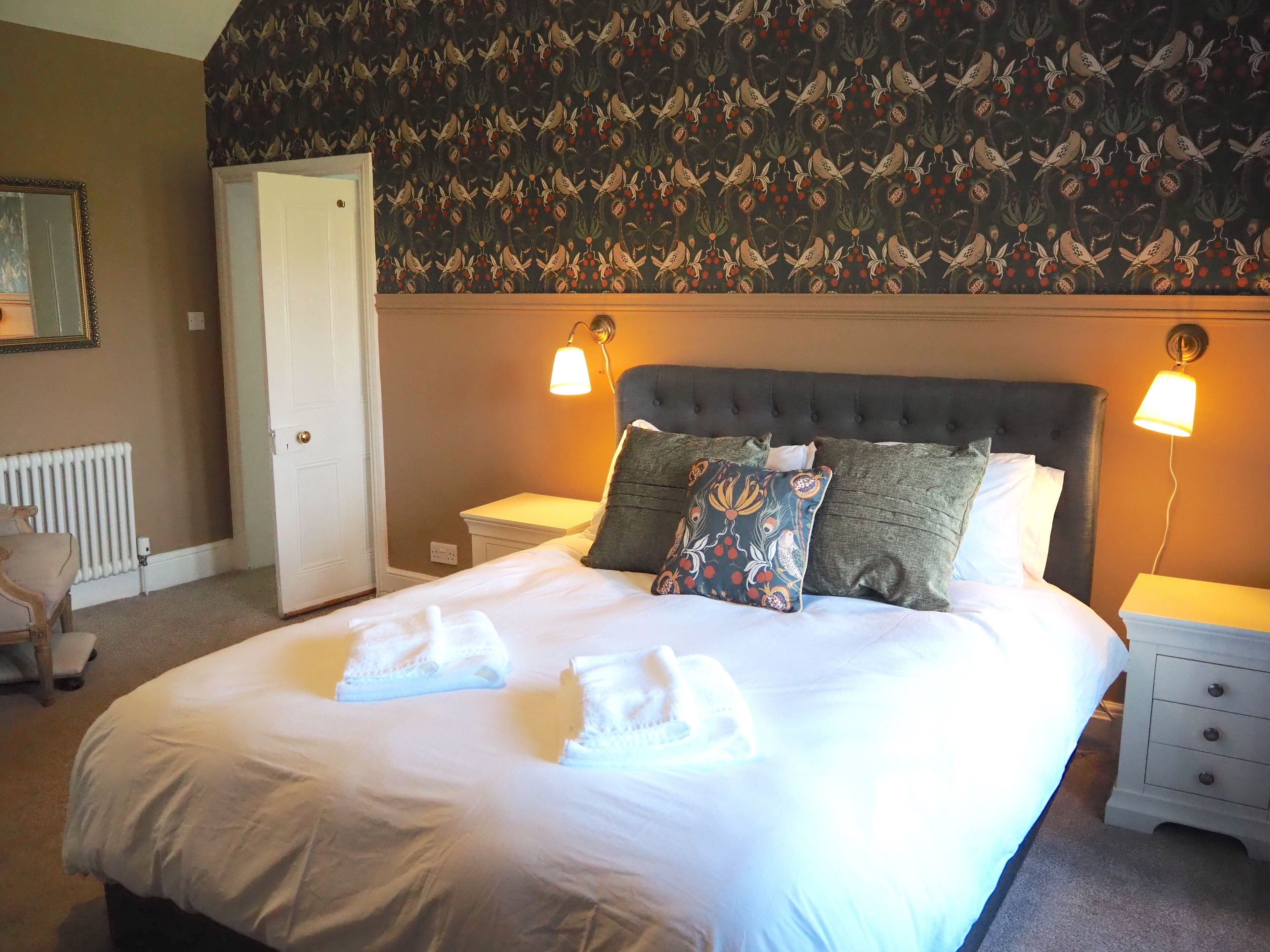 A neatly made bed with white linens and decorative pillows, a bedside table with a lamp, and a patterned wallpaper.