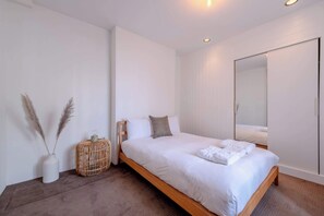 Um quarto moderno com cama de madeira, roupa de cama branca, mesa de cabeceira de vime, um vaso branco com plumas de cortiça e um espelho grande.
