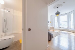 A modern bathroom with a shower, toilet, and a white door leading to a living area with a sofa, a table, and a window.