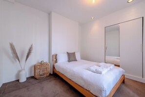 Ein modernes Schlafzimmer mit einem hölzernen Bett, weißer Bettwäsche, einem runden Wickertisch, einer Vase mit Pampasgras und einem großen Spiegel.