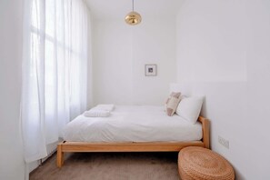 Une chambre à coucher au style minimaliste, avec un lit en bois, du linge de lit blanc, un tabouret en osier et une suspension.