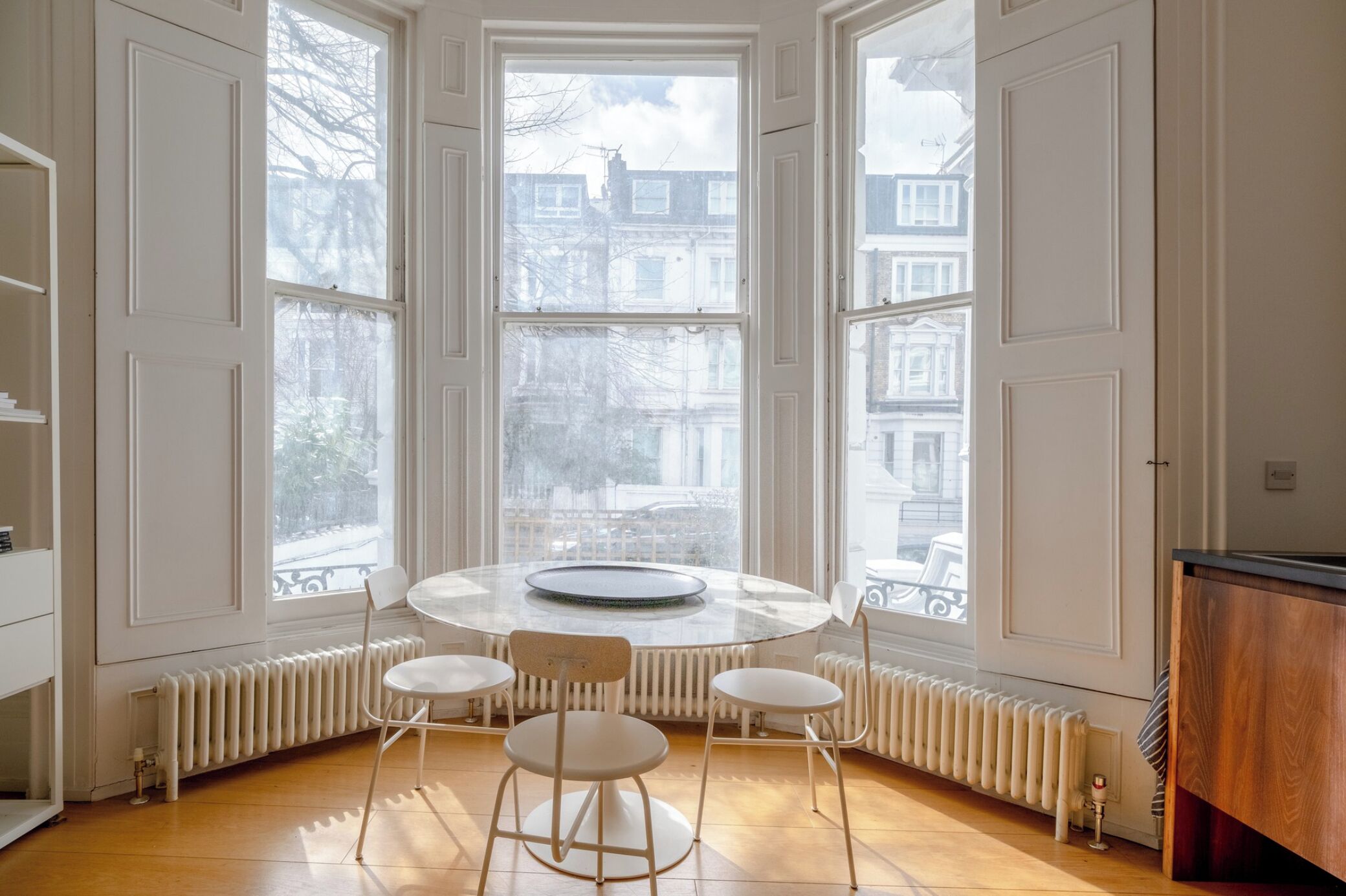 Una habitación con una mesa redonda y cuatro sillas, ventanas amplias con vista a edificios y un radiador debajo de las ventanas.