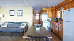 A dining area with a wooden table and chairs, a sofa with decorative pillows, and a kitchen with wooden cabinets and a refrigerator.
