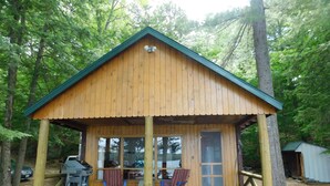 A wooden cabin with a porch, two chairs, a grill, and a small shed in a wooded area.