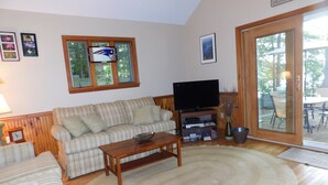 Een woonkamer met een geruite bank, een salontafel, een televisie op een standaard en een groot raam met uitzicht op de omgeving.