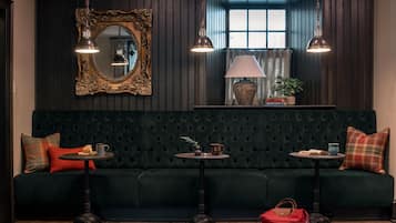 Un sofá tapizado en verde oscuro con respaldo capitonado, dos mesitas redondas con lámparas, un espejo grande y decorativo, y una ventana con cortinas.