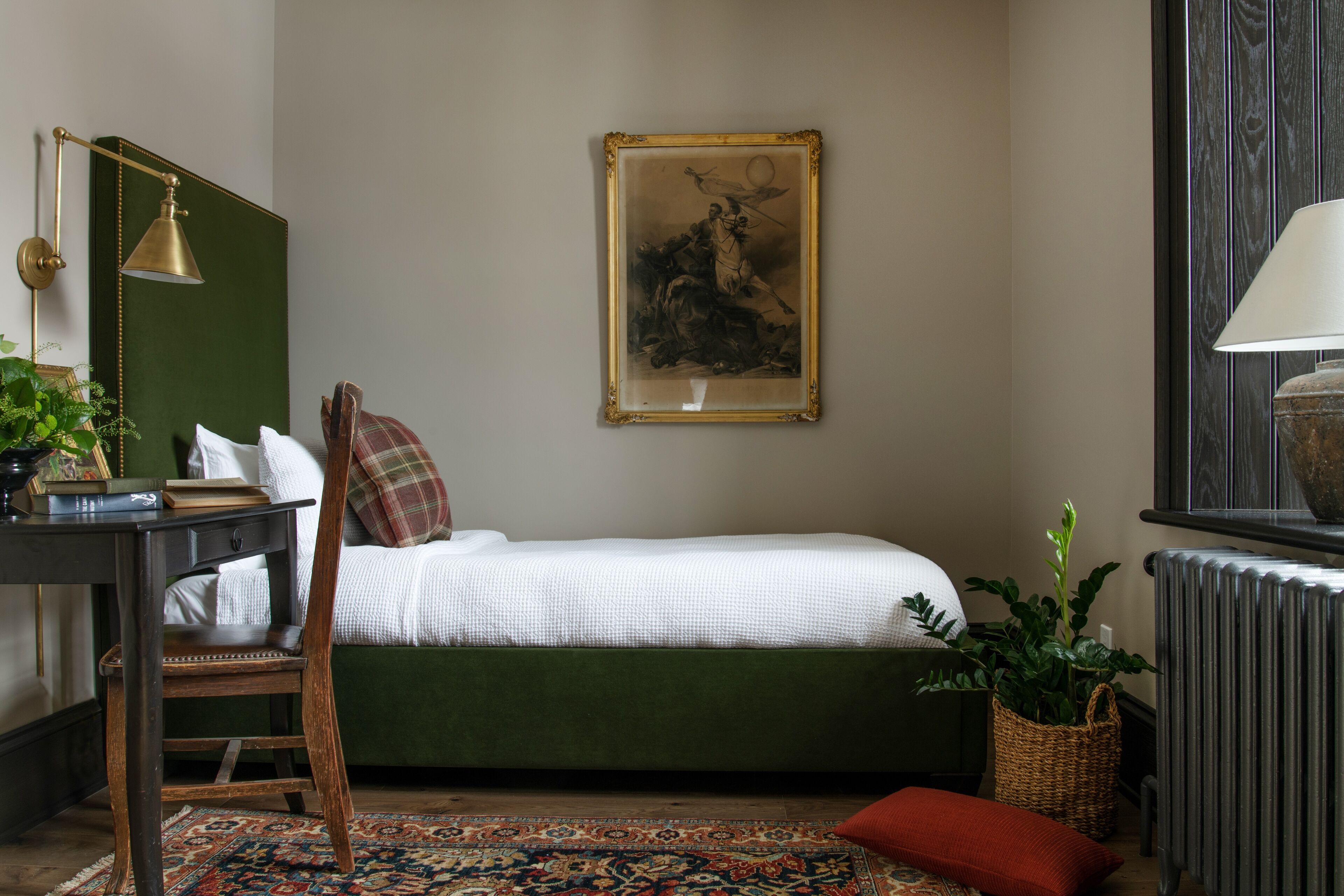 Une chambre avec un lit vert, un bureau avec une lampe, un tableau encadré au mur et un radiateur.