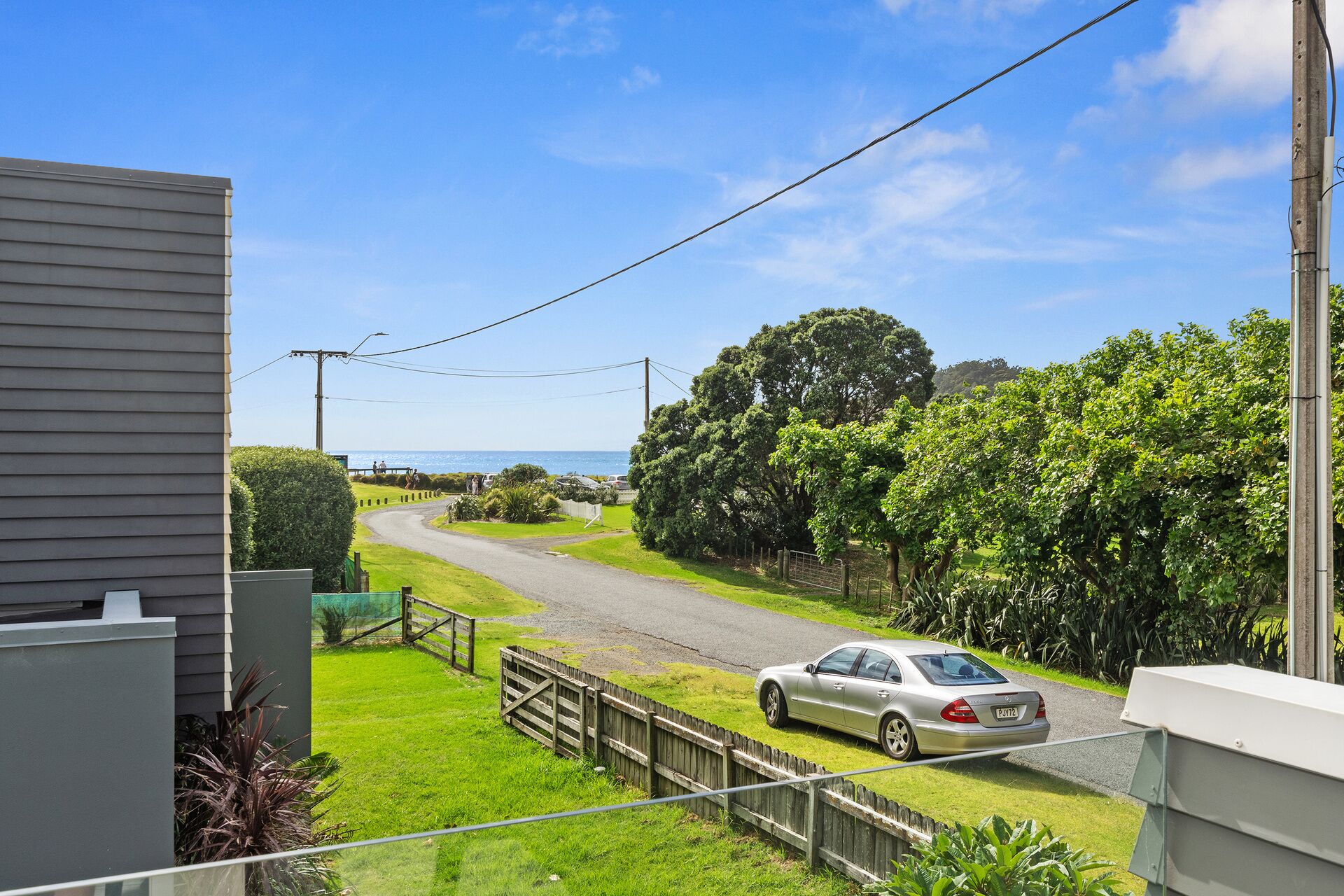 A residential street with a parked car, a wooden fence, and a view of the sea.