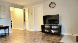 Un soggiorno con pavimento in legno, un mobile porta TV nero, una televisione e una sveglia a parete.