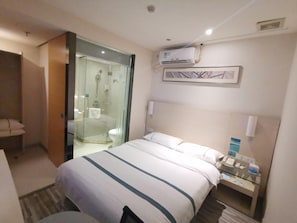 Une chambre d’hôtel comprenant un lit, une douche séparée par des parois en verre, une table de chevet avec un téléphone et un appareil de climatisation fixé au mur.