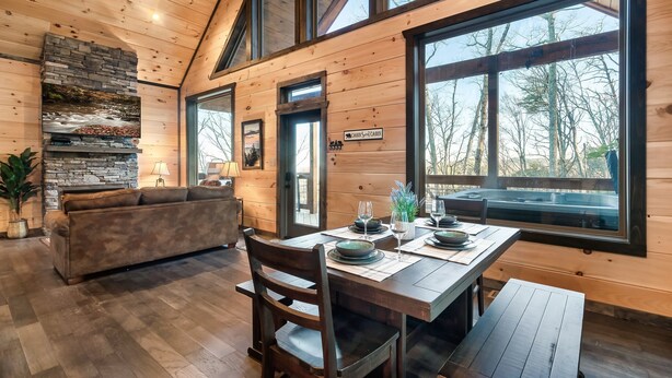 Ein Blockhaus-Interieur mit einem für zwei Personen gedeckten Esstisch, einem Kamin aus Stein und großen Fenstern mit Blick auf den Wald.