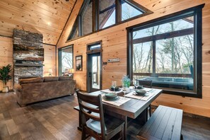 A cabin interior with a dining table set for two, a stone fireplace, and large windows offering a view of the forest. - NEW MTN Retreat ~ Prime Location (Sevierville)