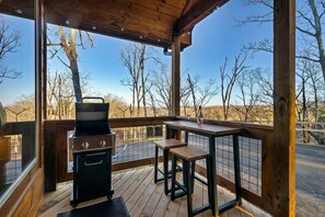 A covered outdoor area with a grill, wooden table, and chairs, overlooking a road and trees. - NEW MTN Retreat ~ Prime Location (Sevierville)
