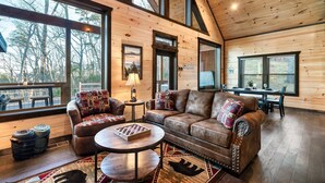 Ein gemütliches Blockhaus-Interieur mit einer großen Holzbalkendecke, einem Ledersofa, einem runden Couchtisch und Blick ins Freie durch große Fenster.