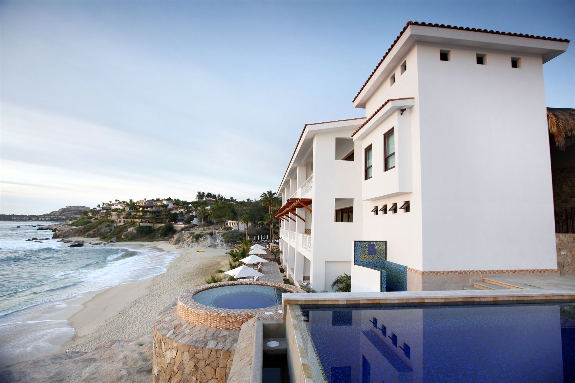 Un hôtel en bord de mer, doté d’une piscine, offrant une vue sur la plage et situé au pied d’une colline où se trouvent des maisons.