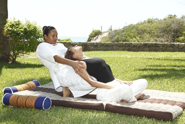 Two people practicing yoga on a mat outdoors with grass and a stone wall in the background.