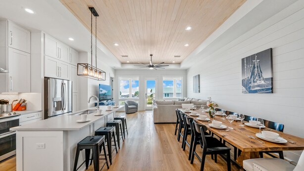 A spacious kitchen with a central island, modern appliances, and a dining area with a wooden table set for six. - One Too Many: 5BR Home on SGI- Beach Views, Priv. Pool, Sleeps 14 (St. George Island)