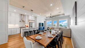 A modern kitchen with white cabinetry, a wooden dining table set for six, and a large island with bar stools. - One Too Many: 5BR Home on SGI- Beach Views, Priv. Pool, Sleeps 14 (St. George Island)