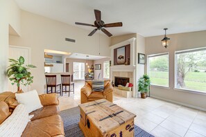 A spacious living room with a leather sofa, a wooden coffee table, and a fireplace. There is a ceiling fan, a dining area in the background, and large windows with a view of the outdoors.