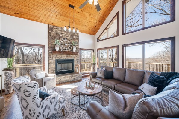 A spacious living room with a stone fireplace, large windows, and a high ceiling.