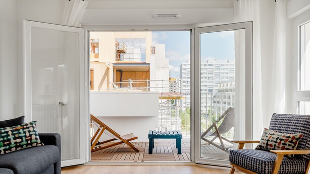 A modern living room with a grey sofa, a patterned armchair, and a wooden bench. A large sliding glass door leads to an outdoor balcony with a view of a cityscape and construction site. - Edgar Suites Meudon-La-Forêt (Meudon)