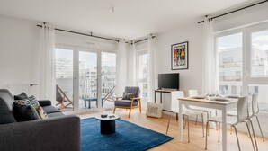 A modern living room with a sofa, a coffee table, a dining table with chairs, a TV, and large windows with curtains. - Edgar Suites Meudon-La-Forêt (Meudon)