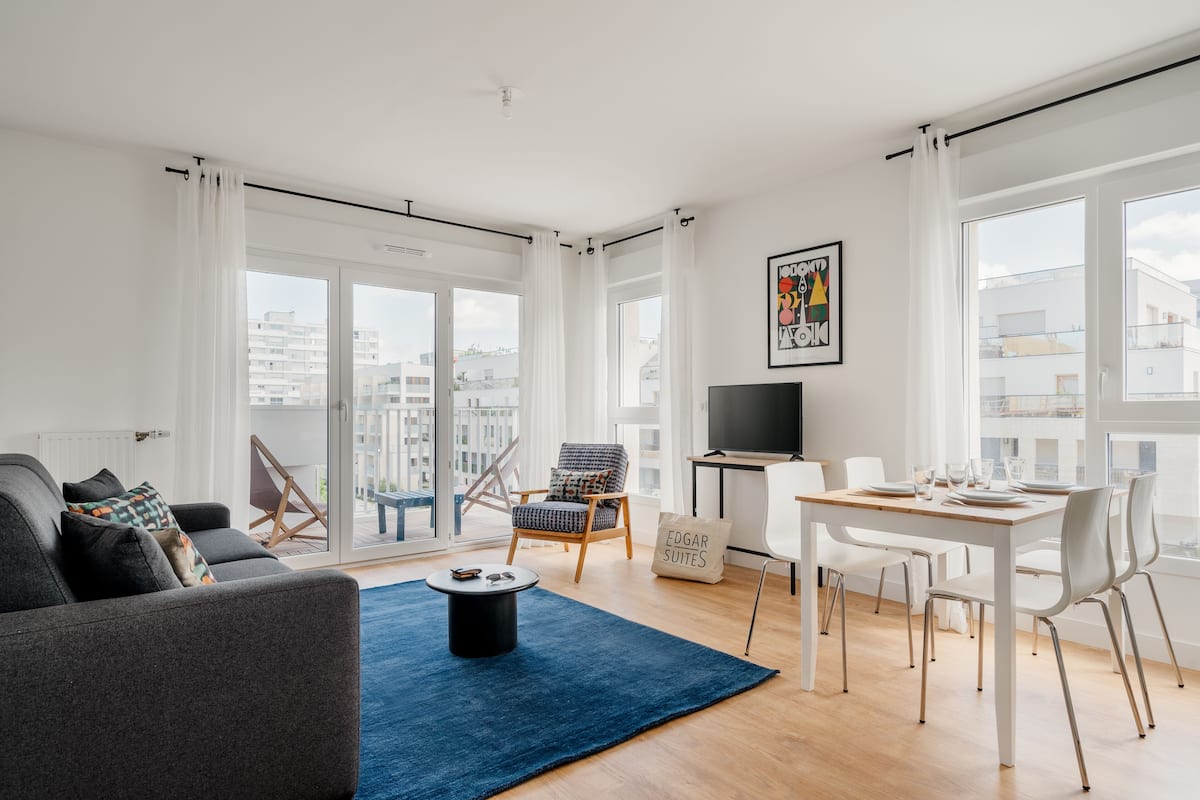 Una sala de estar moderna con sofá, una mesa de centro, una mesa de comedor con sillas, televisor y ventanas grandes con cortinas.