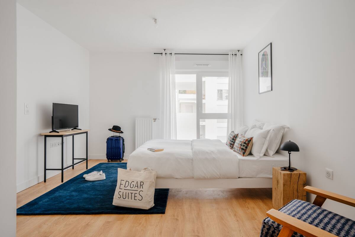 Una habitación de hotel moderna con una cama, un escritorio con televisor, una maleta y un bolso con la etiqueta 'Edgar Suites'.