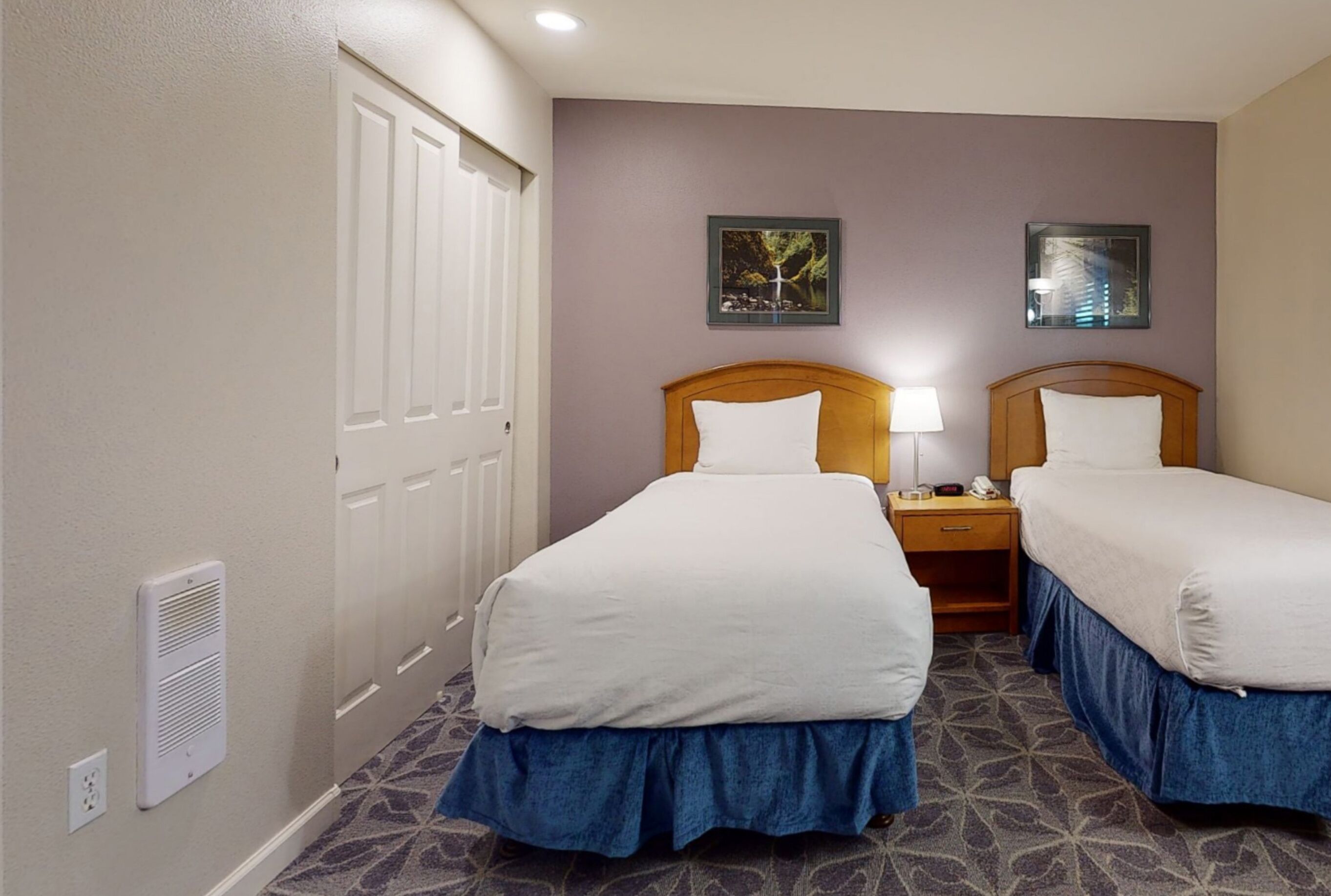 A hotel room with two beds, a wooden headboard, a nightstand with a lamp, and a wall-mounted heater.
