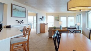 A spacious living area with a wooden dining table, a sofa, and a TV mounted on the wall. There is a fireplace, a large window, and a lamp on the table. - Whale Pointe - Ocean Front 3bd3ba - Assignment 3 (Depoe Bay)