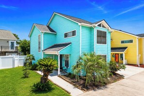 A two-story house with a front porch, a small garden with palm trees, and a neighboring yellow house. - ONE More Happy Day-3BR/3BA Beach House-Sleeps 11 (North Myrtle Beach)