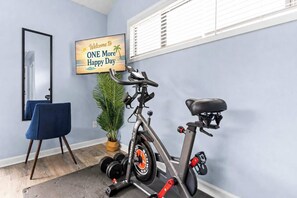 A room with a stationary bike, a mirror, a blue chair, a potted plant, and a welcome sign.
