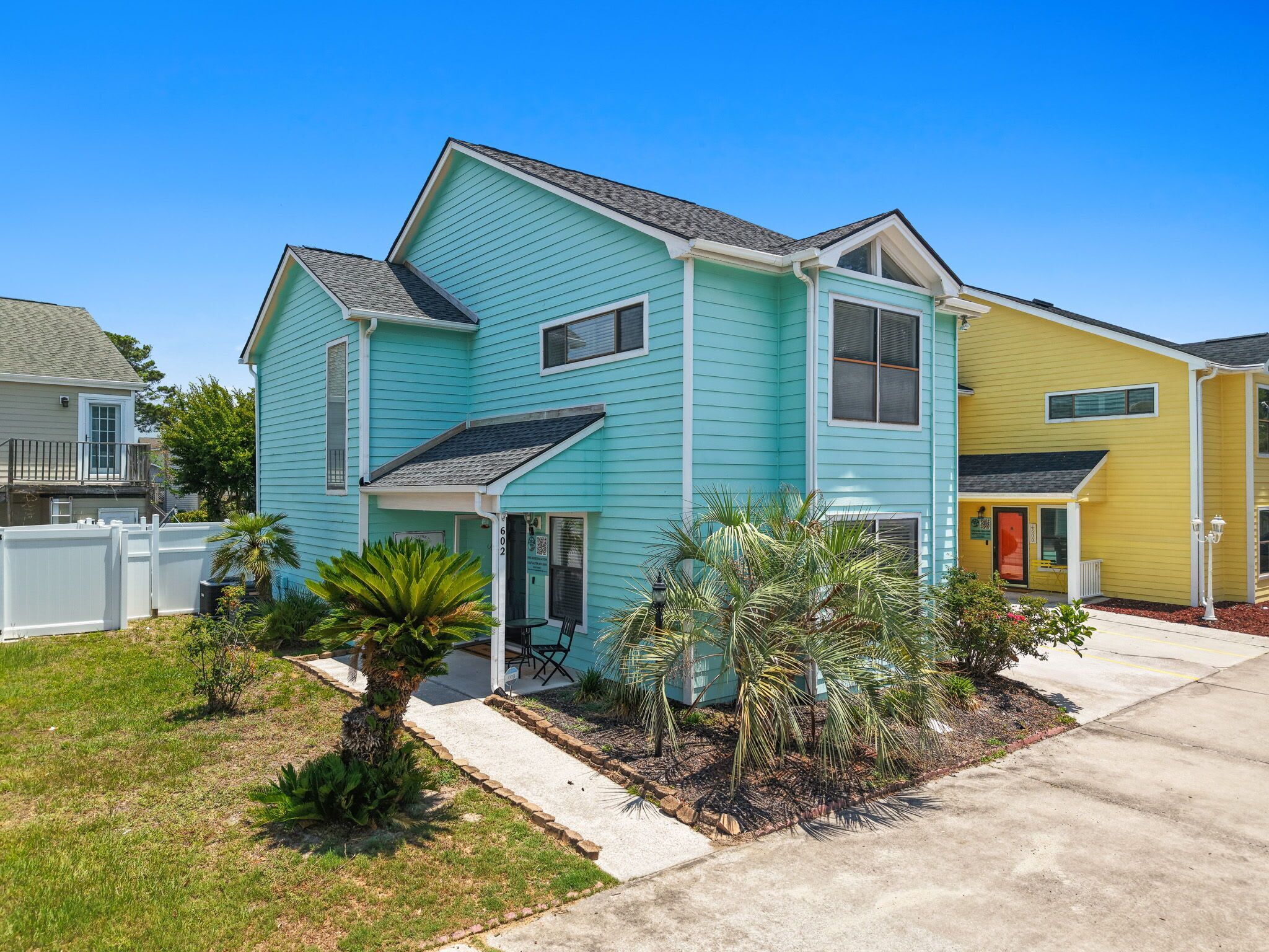 A two-story house with a front porch, a small garden with palm trees, and a neighboring yellow house.