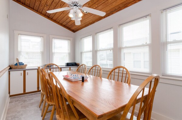 A dining area with a wooden table and chairs, a ceiling fan, and large windows with blinds.