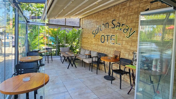 Un espacio exterior moderno con mesas y sillas, una partición de vidrio y una pared de ladrillo con el nombre 'Sultan Saray OTEI'.