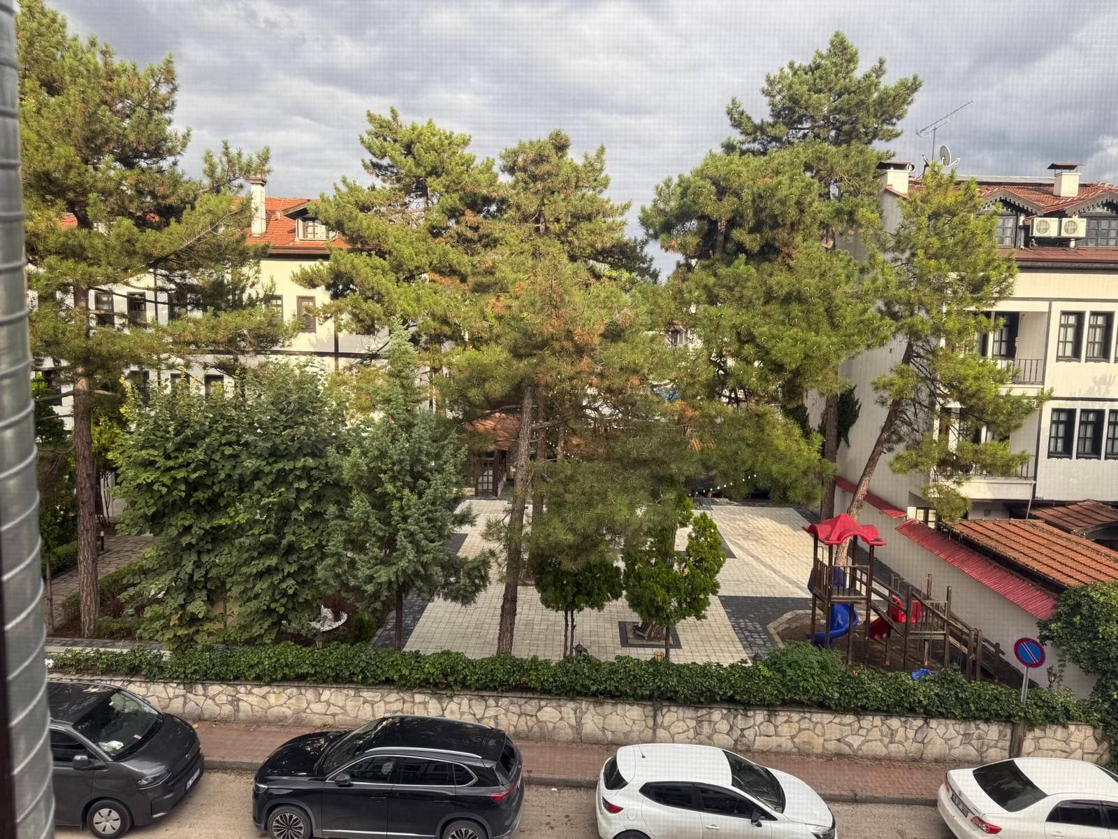 Zona residencial con coches aparcados, árboles y edificios con tejados de tejas rojas.