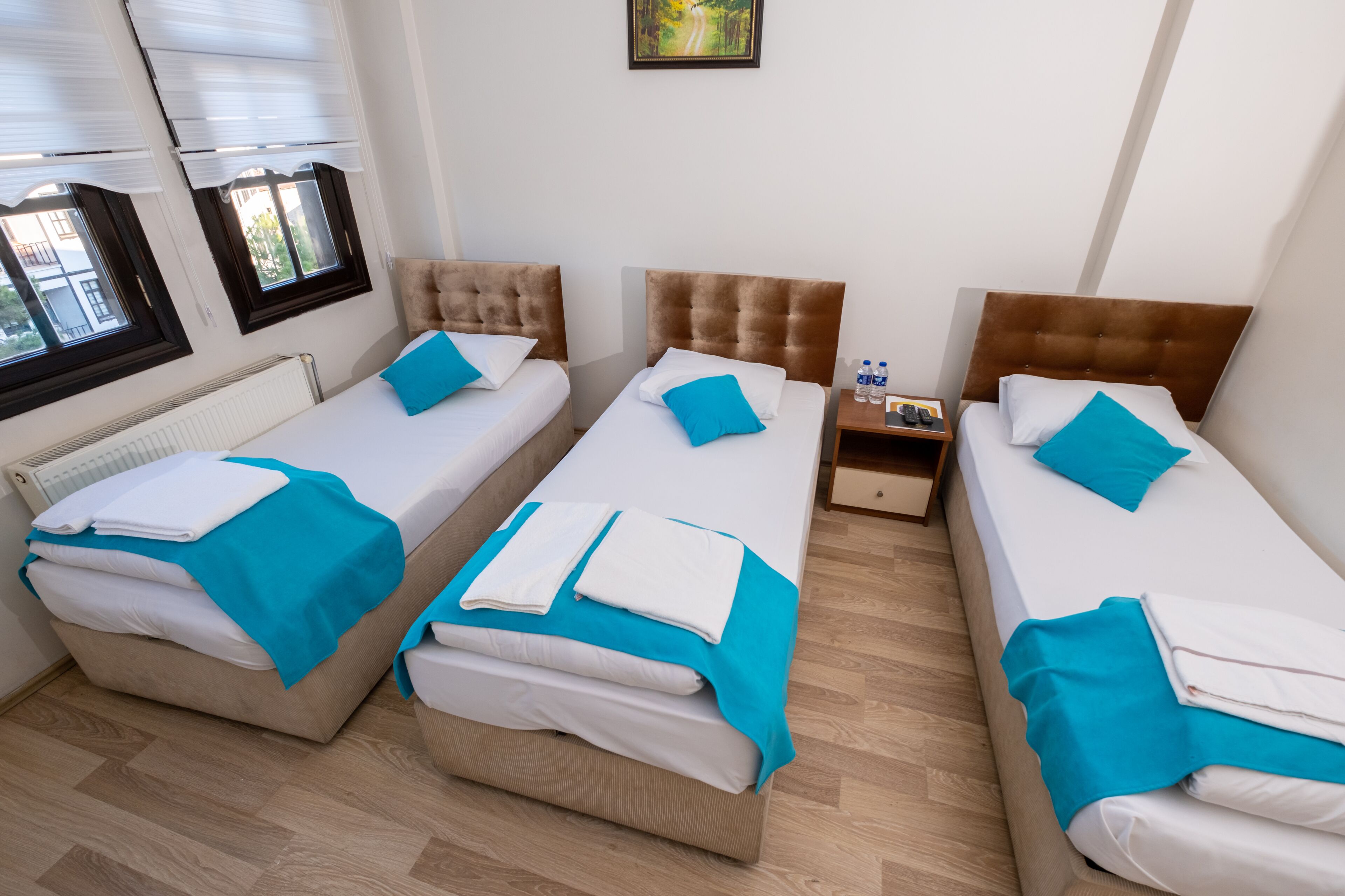 A hotel room with three beds, each with a blue and white bedspread and matching pillows. There is a small bedside table with a lamp and a water bottle.