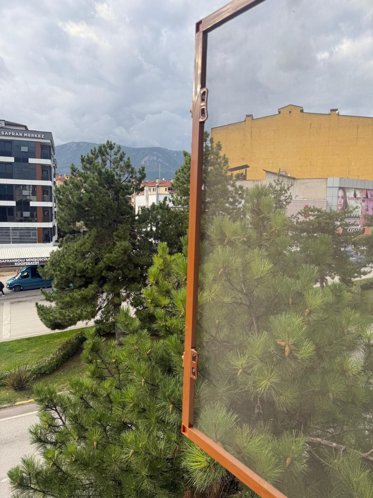 A view through a window with a metal frame, featuring a building with a sign 'SAFRAN MERKEZ' and a mountain in the background.