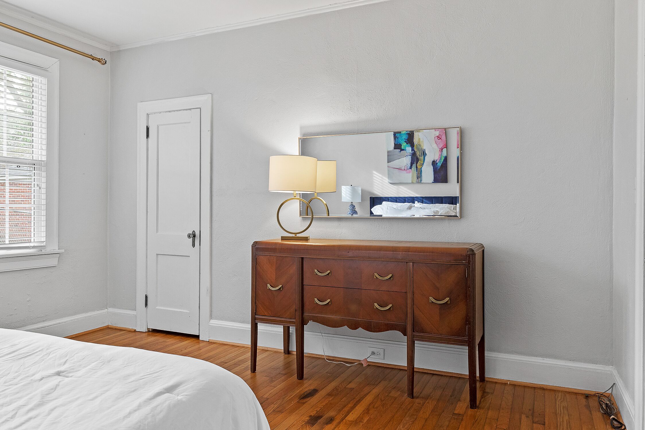 A bedroom with a wooden dresser, a bed, a white door, and a window with blinds.