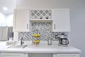 A kitchen with white cabinets, a tiled backsplash, a coffee maker, and a fruit basket.