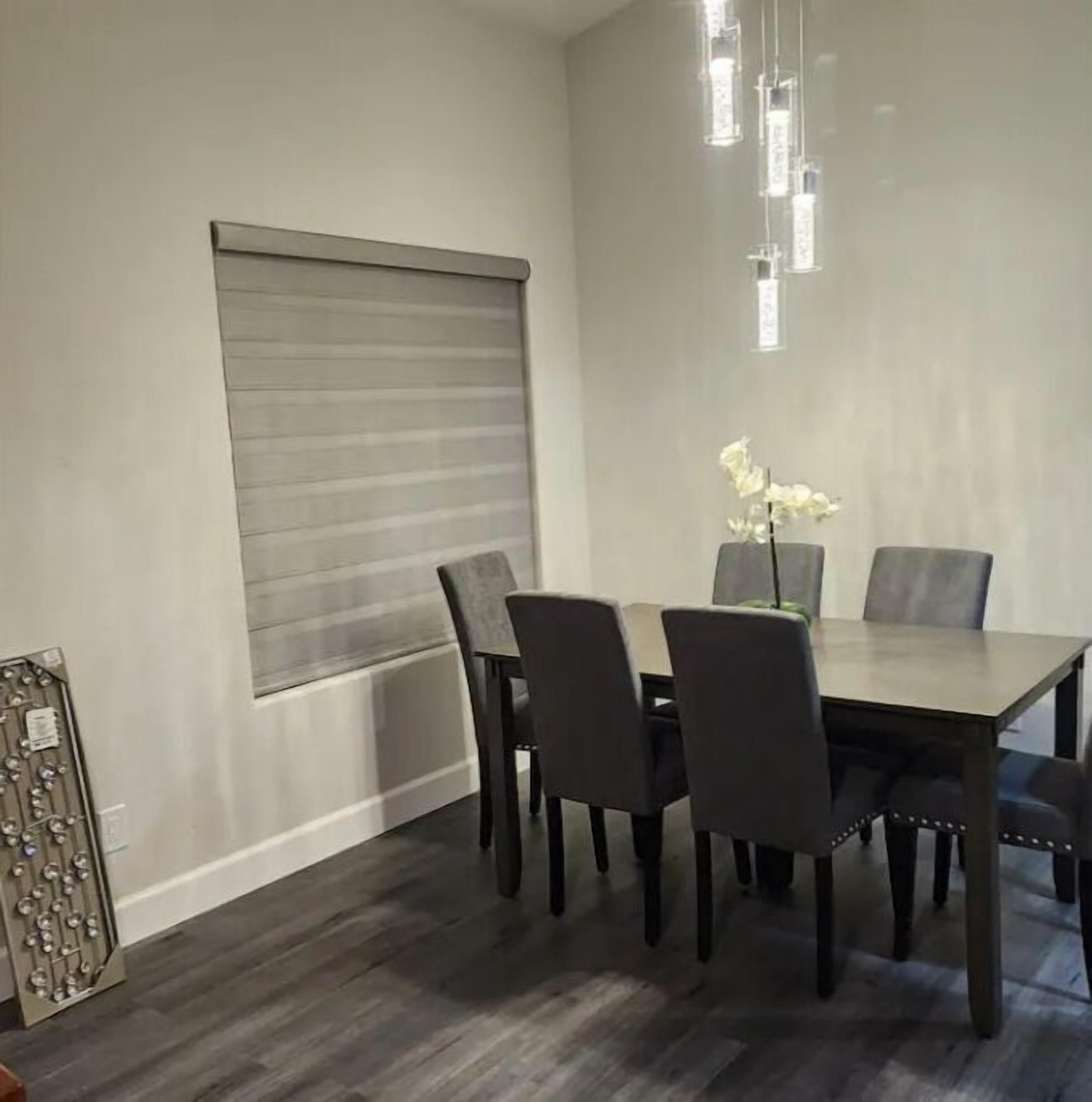 A dining area with a rectangular table, six chairs, a window with a roller blind, and a decorative metal panel on the left.
