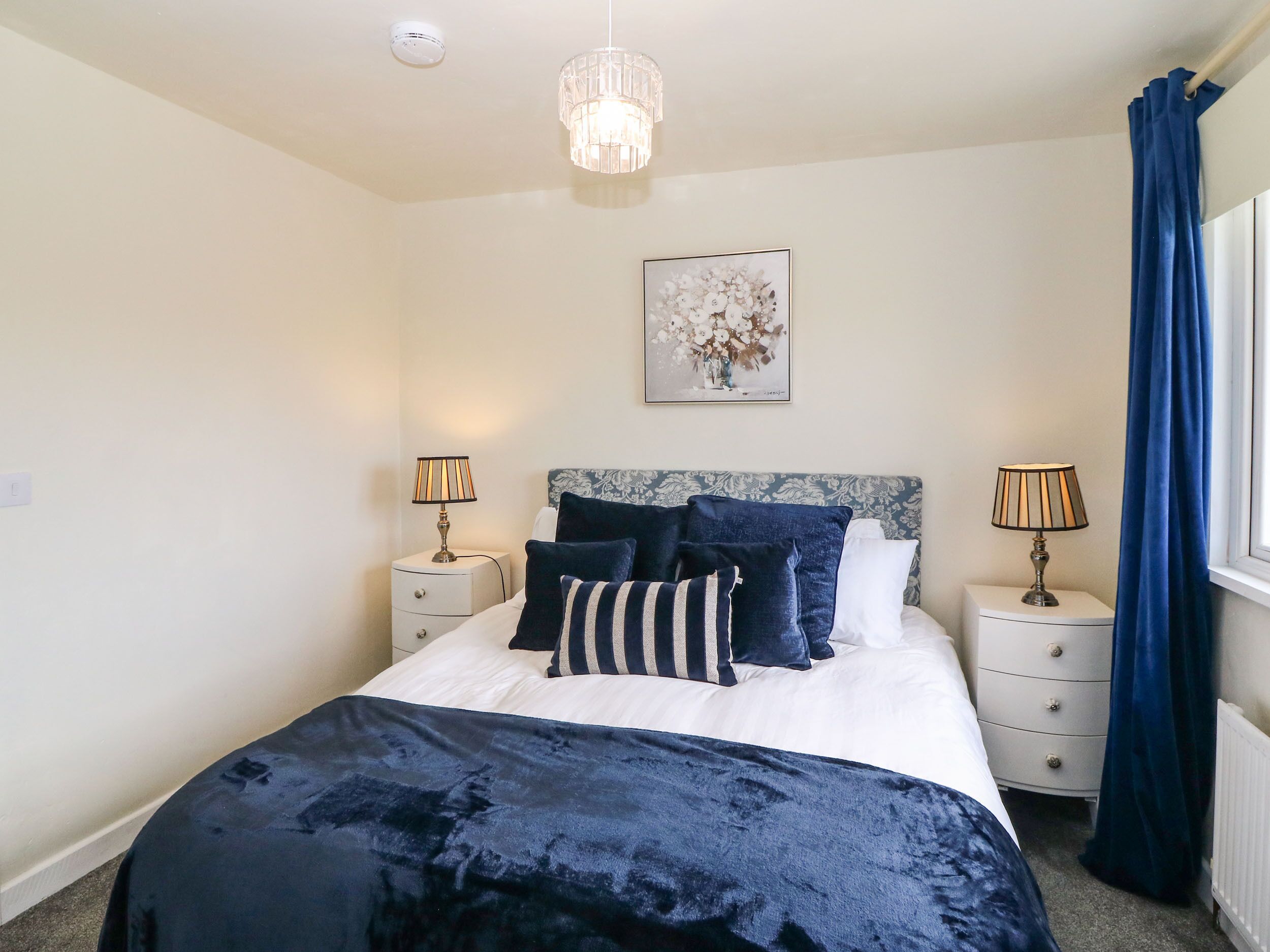 A bedroom with a bed, two bedside tables, a lamp, a ceiling light, a window with curtains, and a wall-mounted artwork.