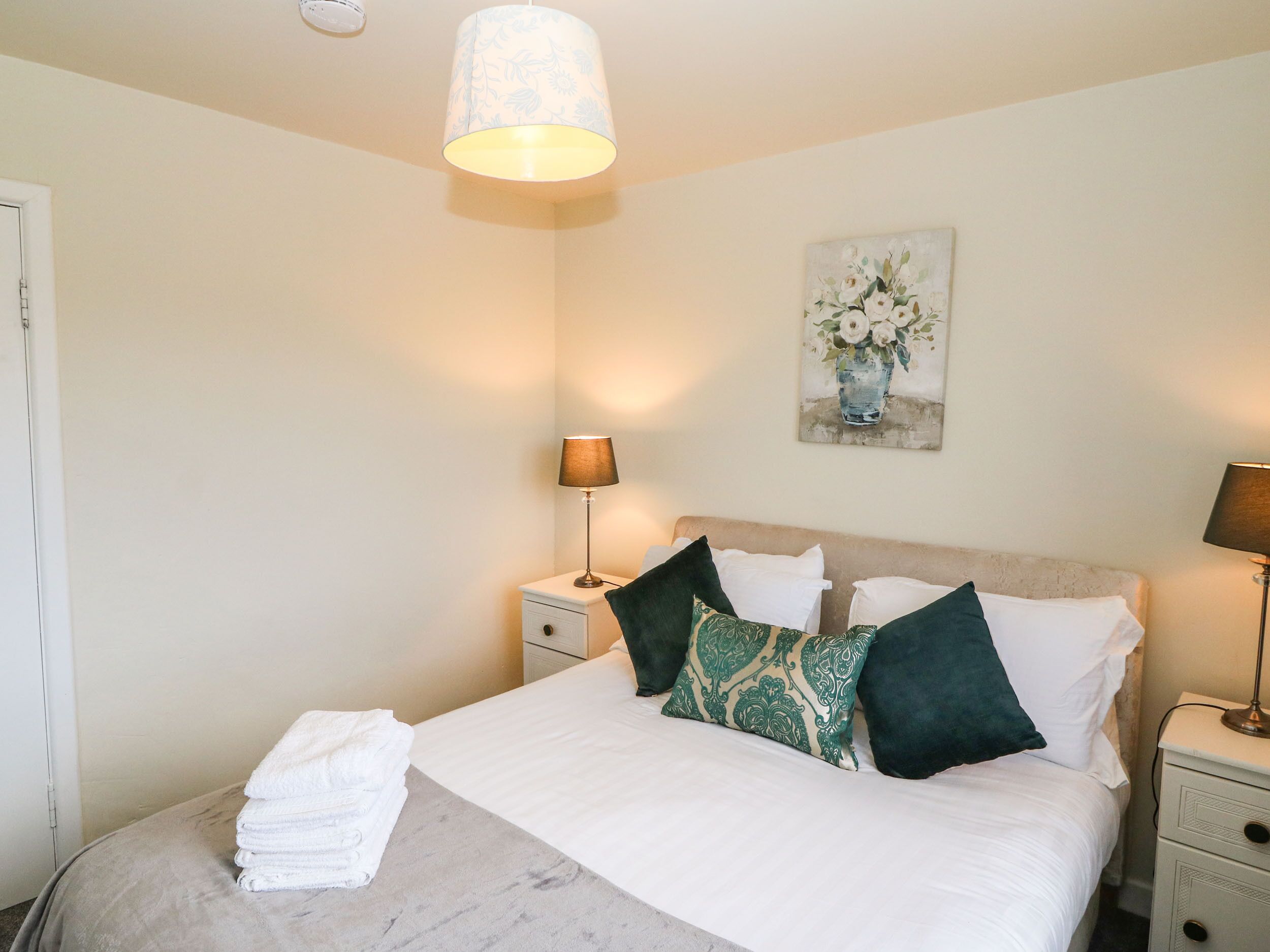 A neatly made bed with white linens and decorative pillows, a bedside lamp, a small bedside table with a lamp, and a wall-mounted painting of flowers.