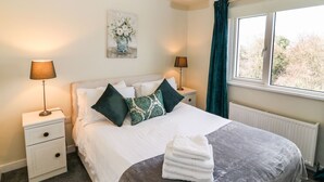 Een netjes opgemaakt bed met een grijze deken, twee nachtkastjes met lampen, een raam met gordijnen, en een schilderij aan de muur.
