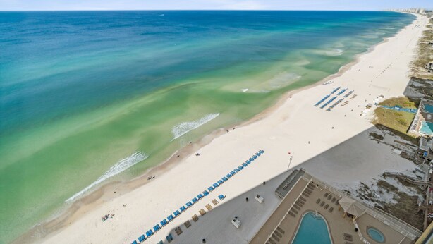 An aerial view of a beach with clear waters, a sandy shore, and a pool area with lounge chairs.
