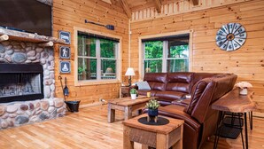 A log cabin interior with a stone fireplace, wooden walls, a leather sofa, a wooden coffee table, and a clock on the wall. - Hidden Creek Lodge by The Inn & Spa at Cedar Falls (Creola)