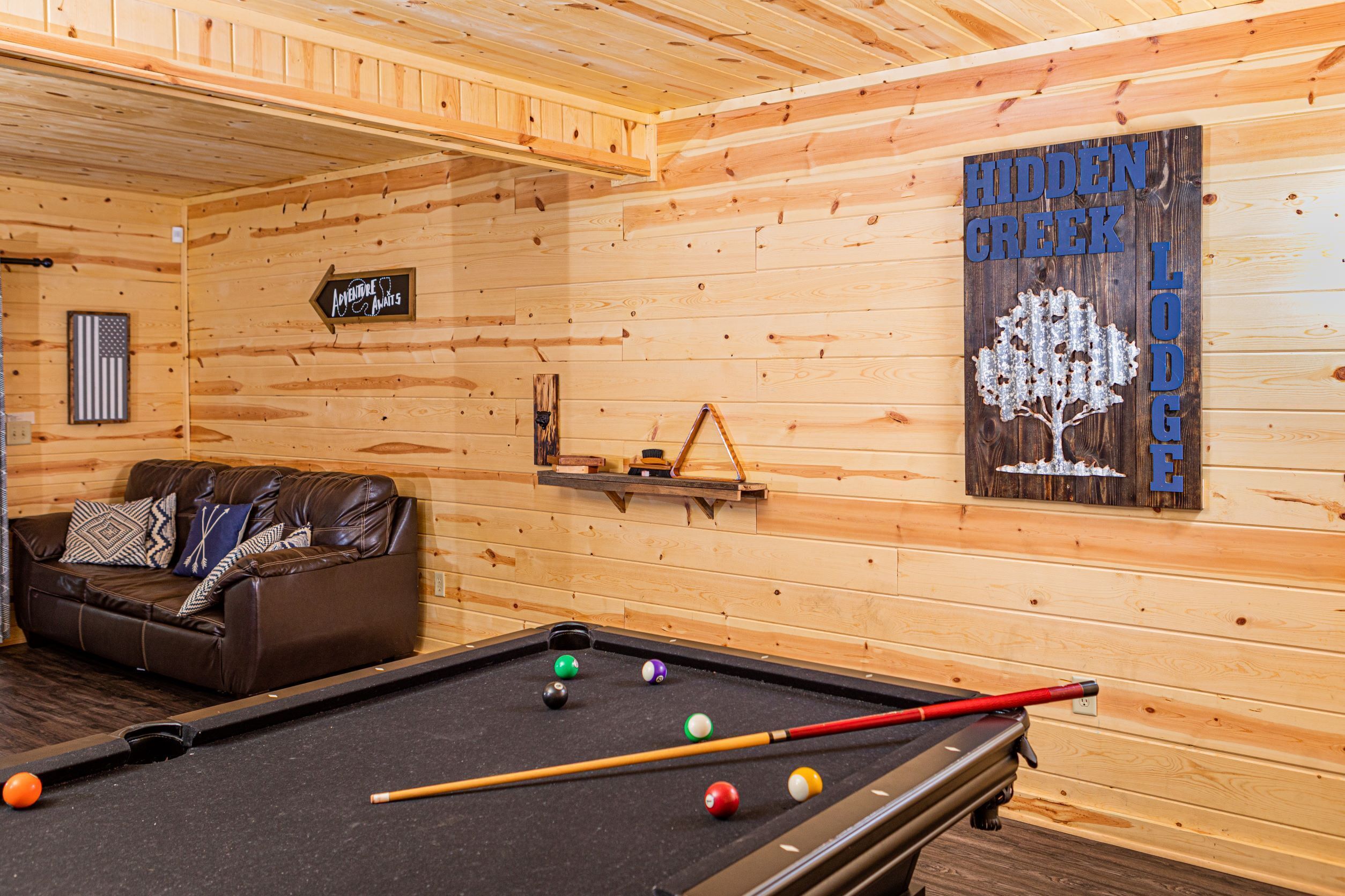 A wooden cabin interior with a pool table, a brown leather sofa, and a sign reading "Hidden Creek Lodge".