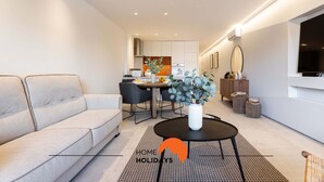 Una sala de estar moderna con un sofá, una mesa de comedor con sillas, una mesa de centro con un florero, y una cocina al fondo.