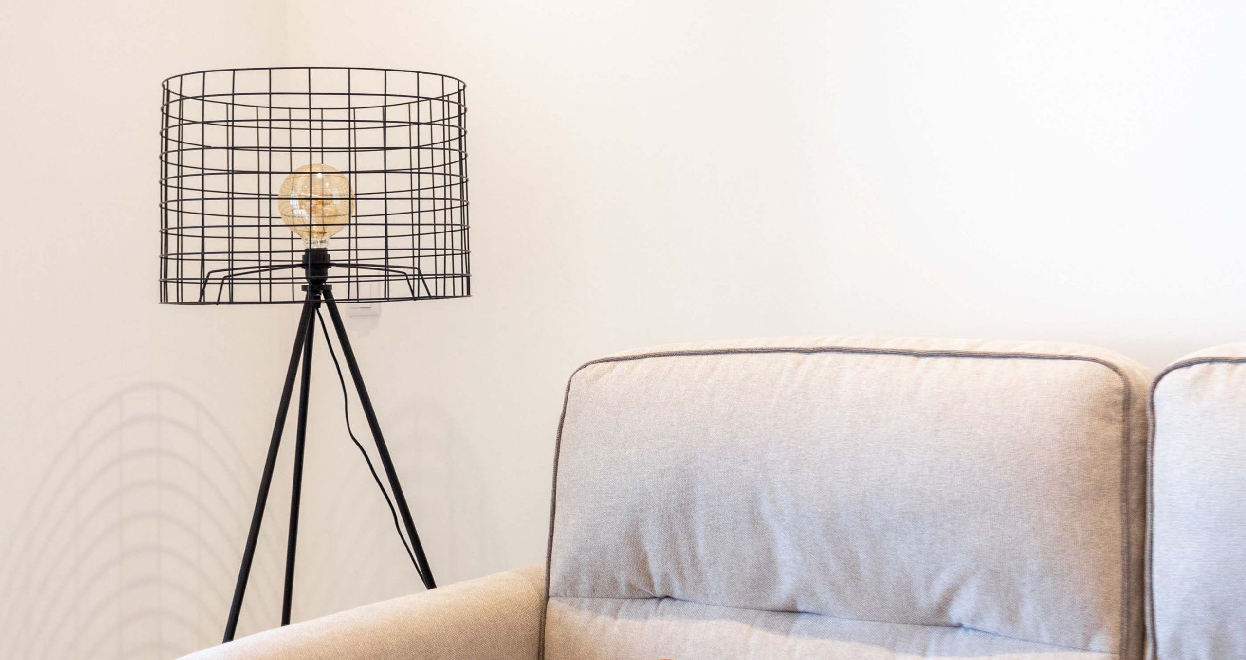 A beige armchair with a cushion, a floor lamp with a wireframe shade, and a wall with a subtle pattern.